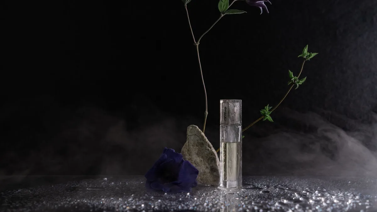 Artistic still life featuring purple flowers, rock, and glass perfume bottle against black background.